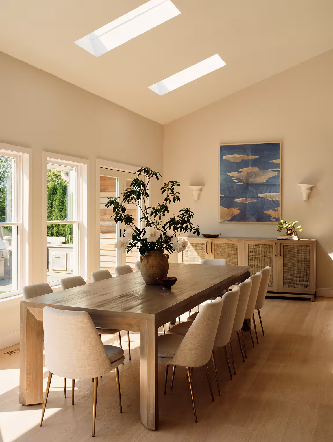 A sunlit dining room with skylights, a long wood table, cream chairs, and a framed blue abstract painting.