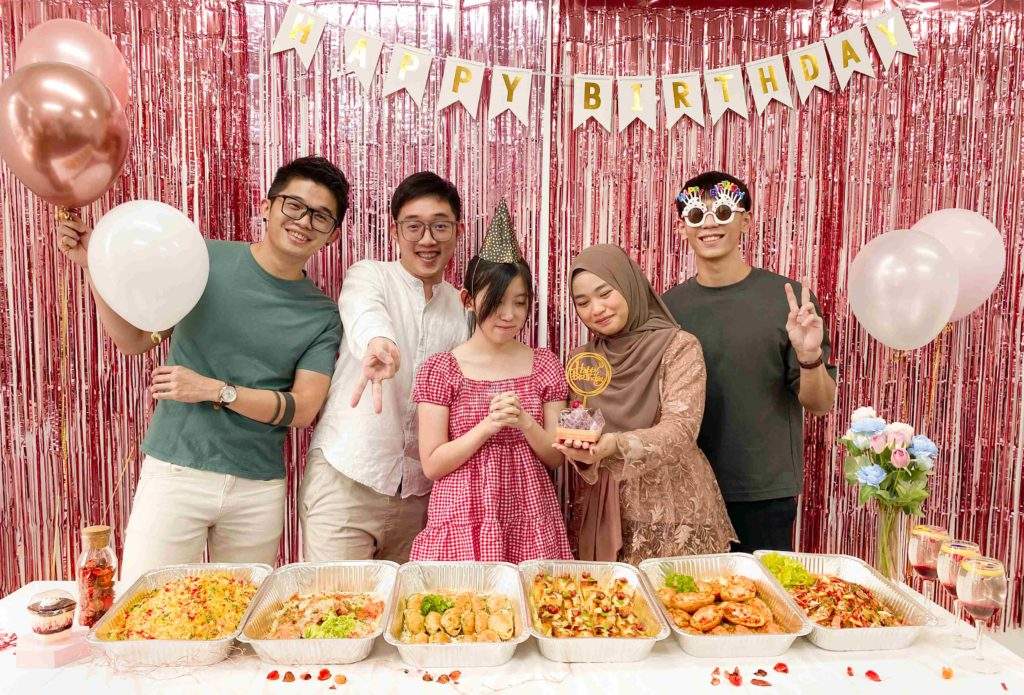 Friends holding a cake behind foil trays of party food at a birthday party catered by iDoCatering, 2022