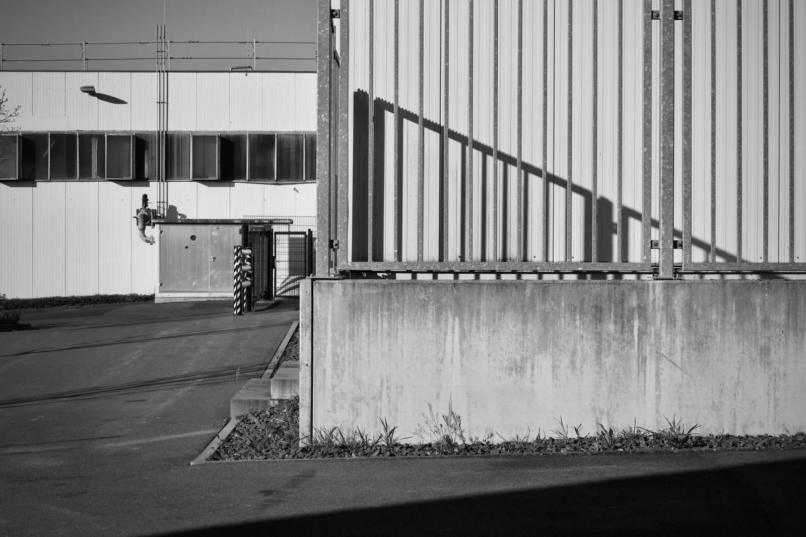 Industrial facade in high-contrast grayscale, a hard diagonal shadow crossing corrugated steel