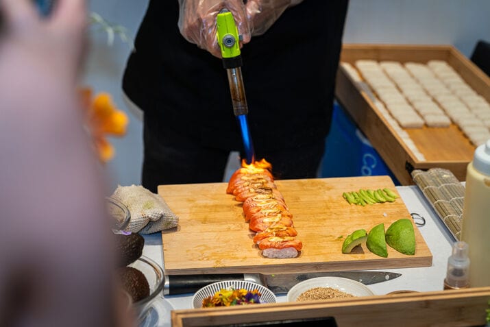 Chef torching aburi salmon at a live sushi station beside sliced avocado — iDoCatering