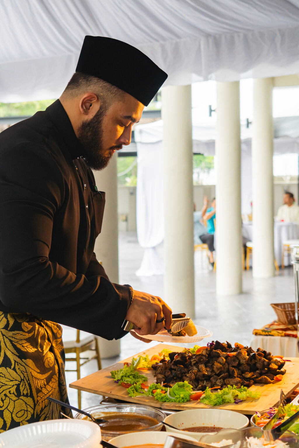 Chef in baju Melayu and songkok plating grilled lamb from a wooden board at an iDoCatering Hari Raya live station