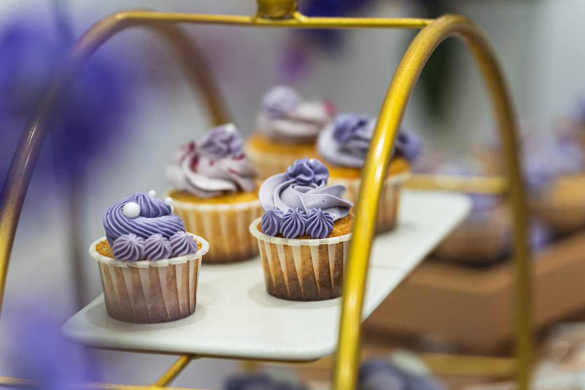 Purple-frosted cupcakes on a gold tiered stand at a lilac-themed party dessert table by iDoCatering