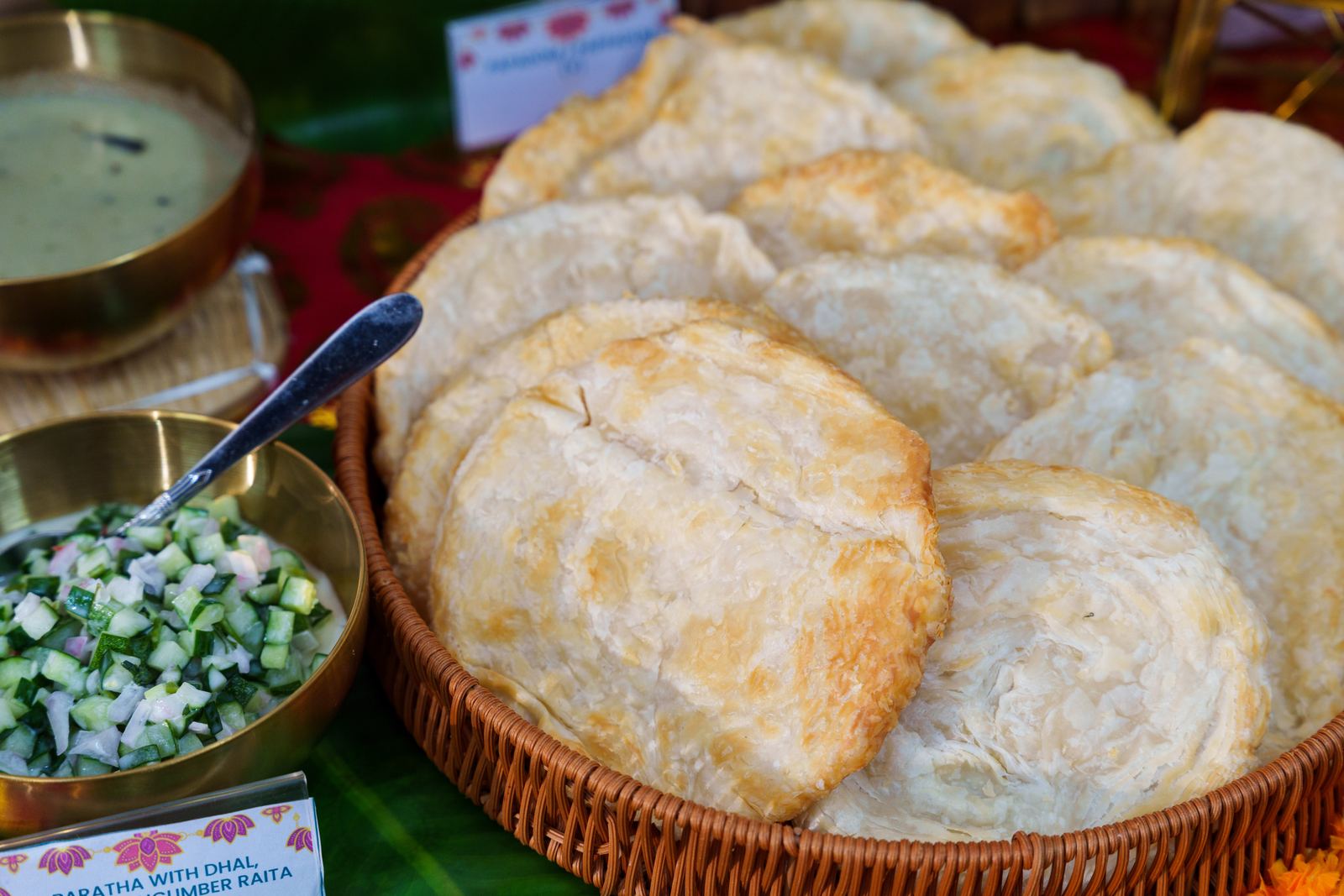 Flaky paratha in a rattan basket with brass bowls of dhal and cucumber raita at a Deepavali buffet