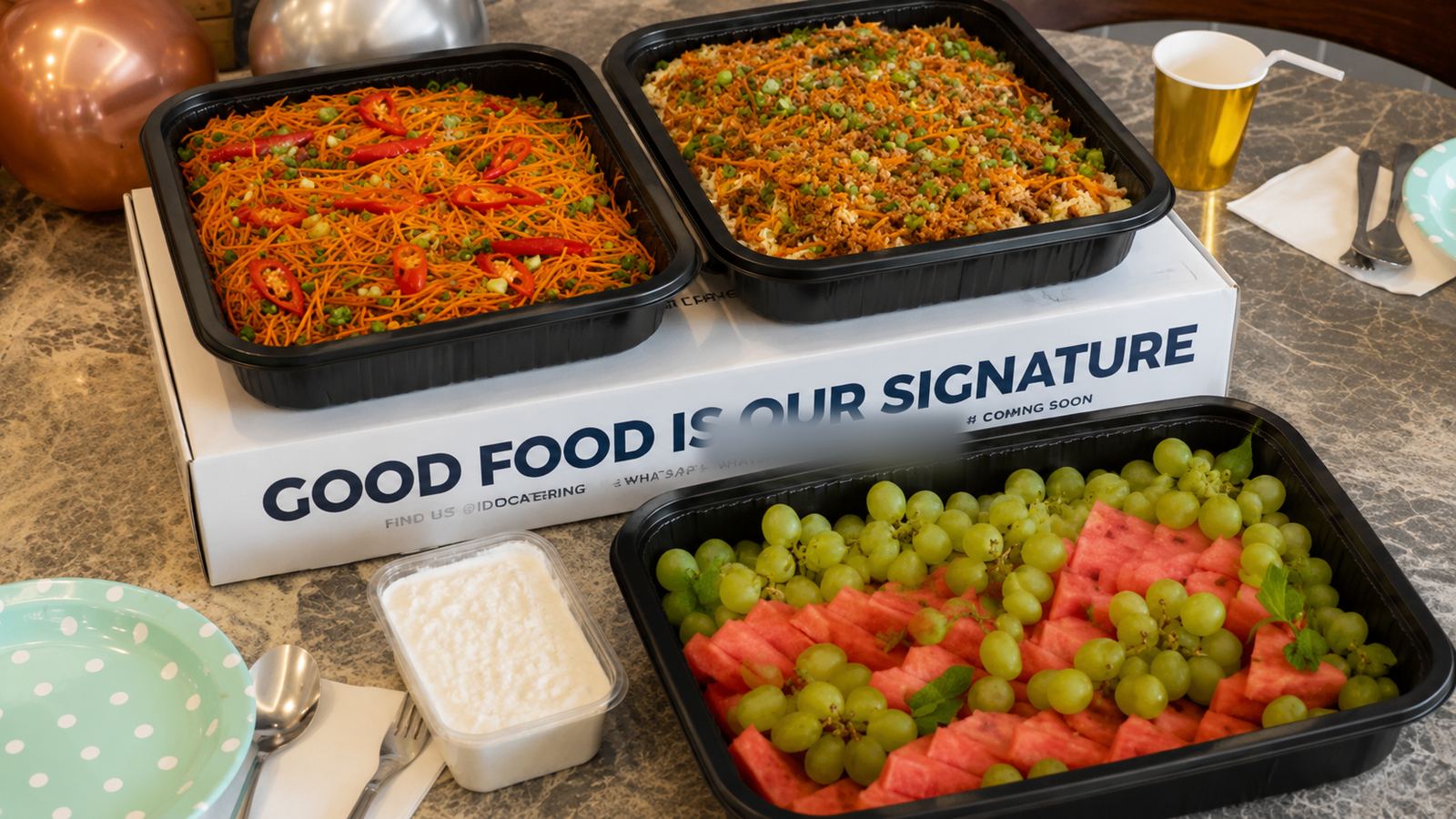 Black catering trays of fried meehoon and fried rice beside a fresh fruit platter of watermelon and grapes
