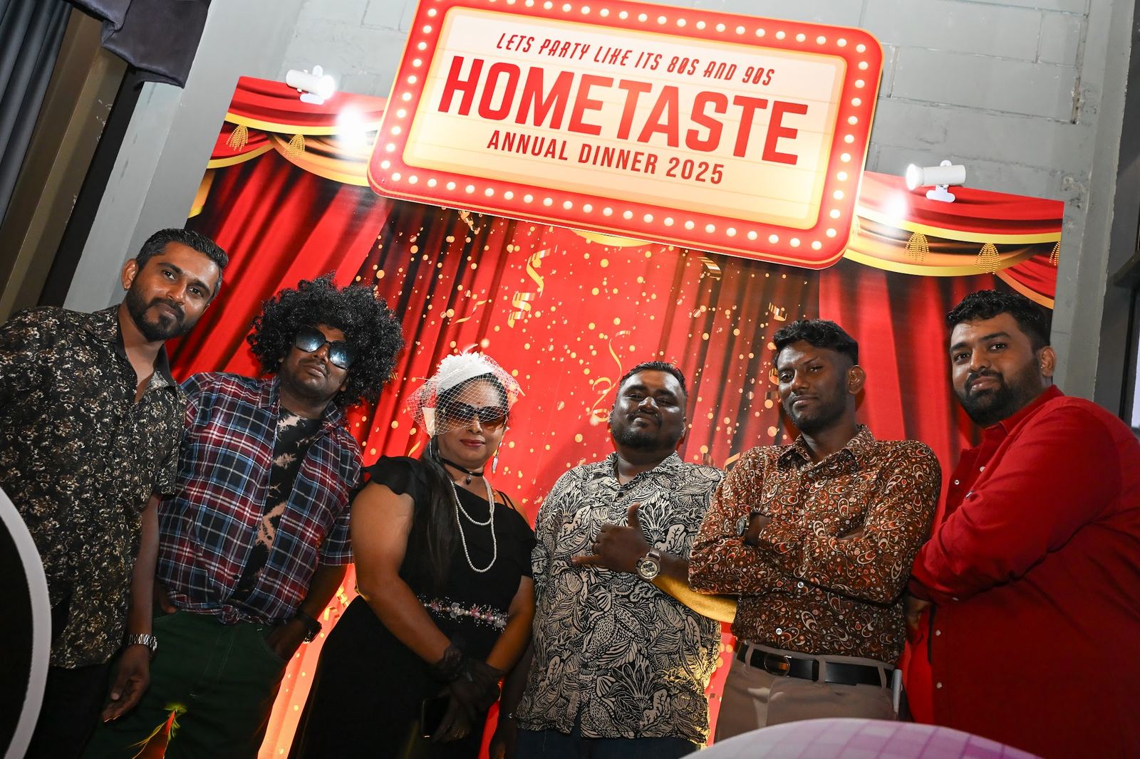 Six colleagues in 80s and 90s outfits at the Hometaste annual dinner 2025 backdrop, a lit marquee sign above them