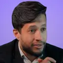 Man with dark hair and beard speaking against a purple-blue gradient background.