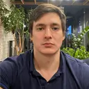 Man with light brown hair wearing a dark blue shirt, sitting indoors with plants in the background.