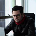 Man with glasses wearing a black jacket and red scarf sitting near a window.