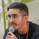 Man with glasses and short hair speaking into a microphone outdoors with blurred greenery background.