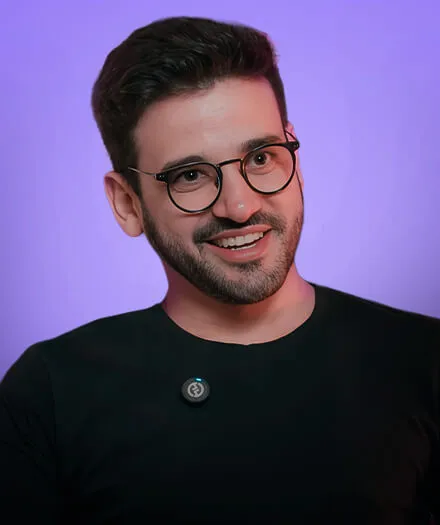 Smiling man with short dark hair and round glasses wearing a black shirt against a purple background.