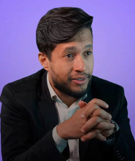 Man with beard and short hair wearing a dark blazer and shirt, hands clasped, against a purple background.