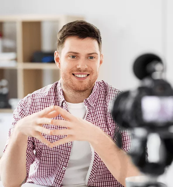 Smiling man in a checkered shirt speaking in front of a camera in a home office.