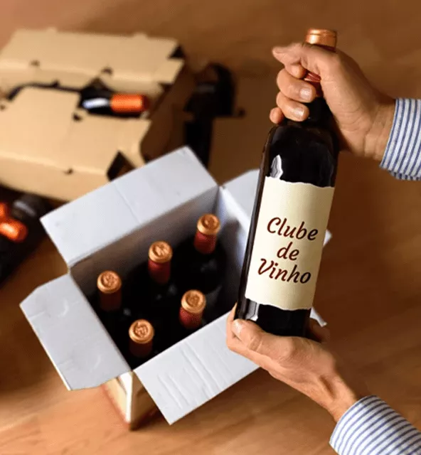 Hands holding a wine bottle labeled 'Clube de Vinho' above an open box with six more bottles inside.