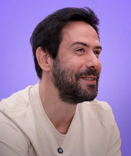 Man with dark hair and beard smiling, wearing a light-colored shirt against a purple background.