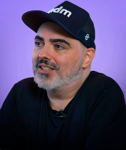 Man with gray beard wearing a black cap and shirt against a purple background.