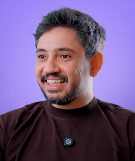Smiling man with gray-streaked hair and beard wearing a dark shirt against a purple background.