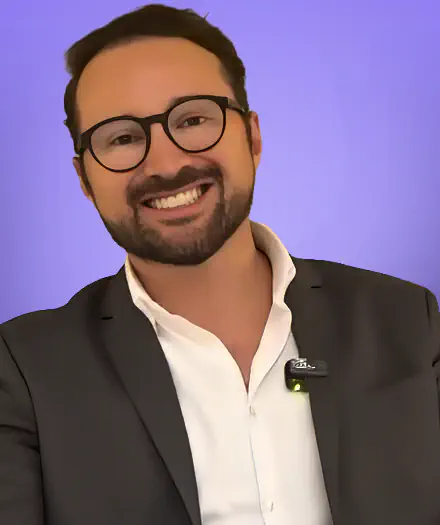 Smiling man with glasses, beard, and dark suit against a purple background.