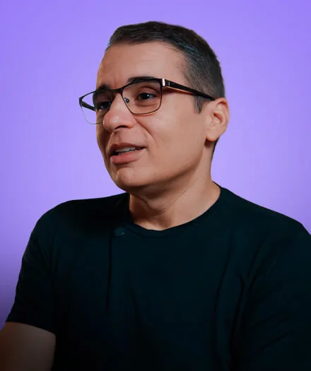 Man wearing glasses and a black shirt against a purple background looking to the side.