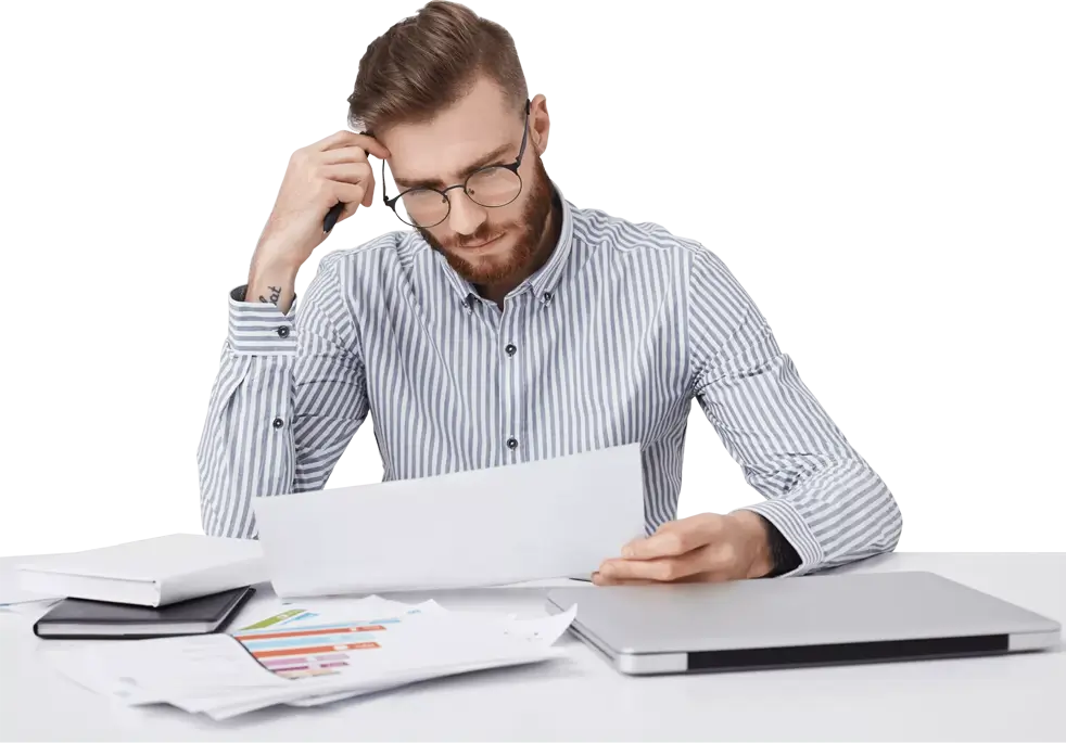 A man with glasses and a striped shirt sits at a table, looking worried and thoughtful at a document in his hands, surrounded by reports and a laptop.