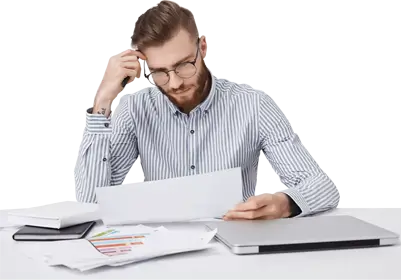 A man with glasses and a striped shirt sits at a table, looking worried and thoughtful at a document in his hands, surrounded by reports and a laptop.