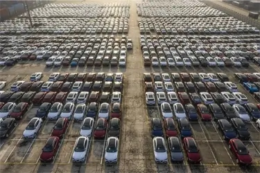 Aerial view of thousands of new cars parked in tight rows at a massive shipping port parking lot.