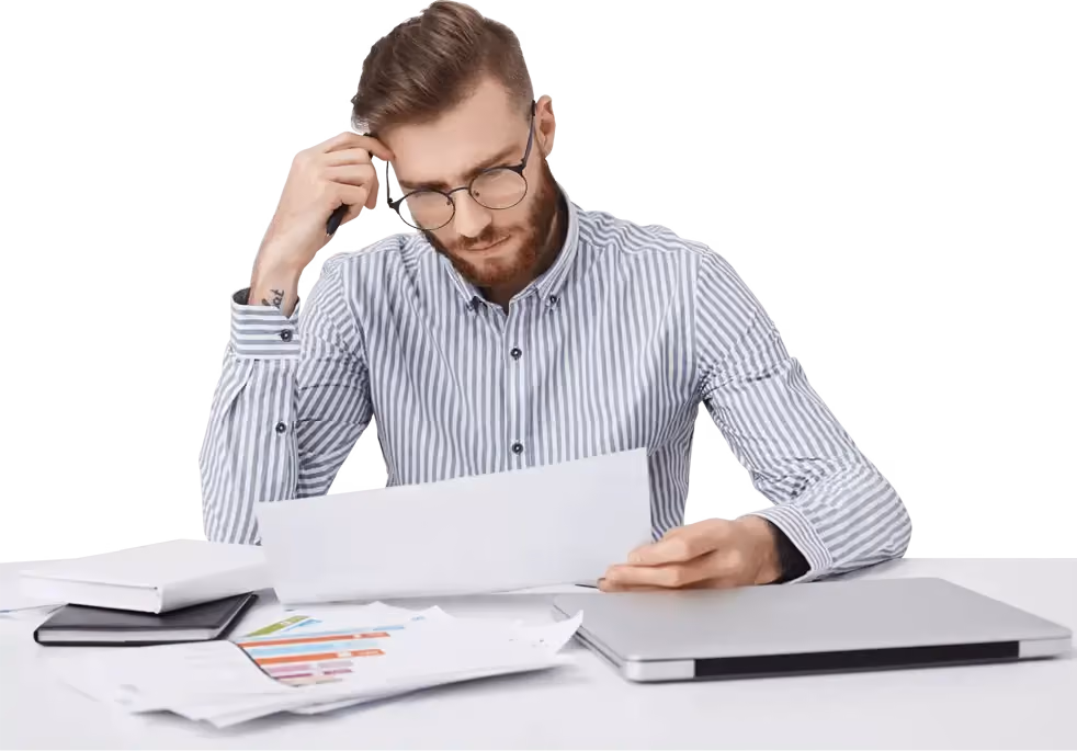 A man with glasses and a striped shirt sits at a table, looking worried and thoughtful at a document in his hands, surrounded by reports and a laptop.