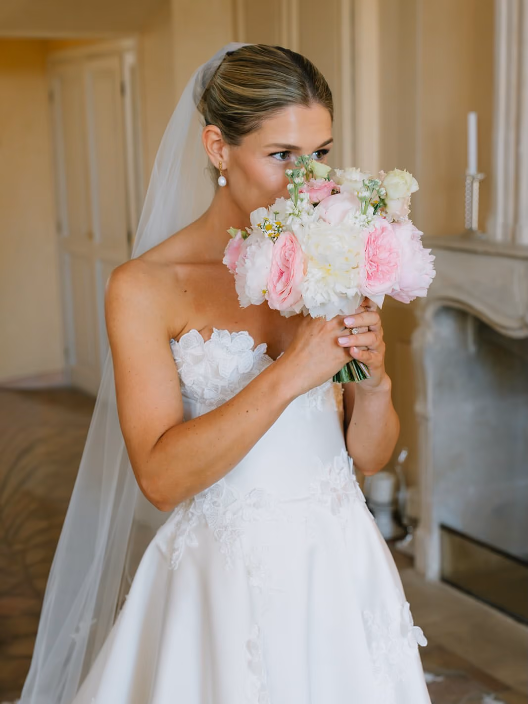 Bride in white strapless dress holding and smelling a pink and white flower bouquet indoors.