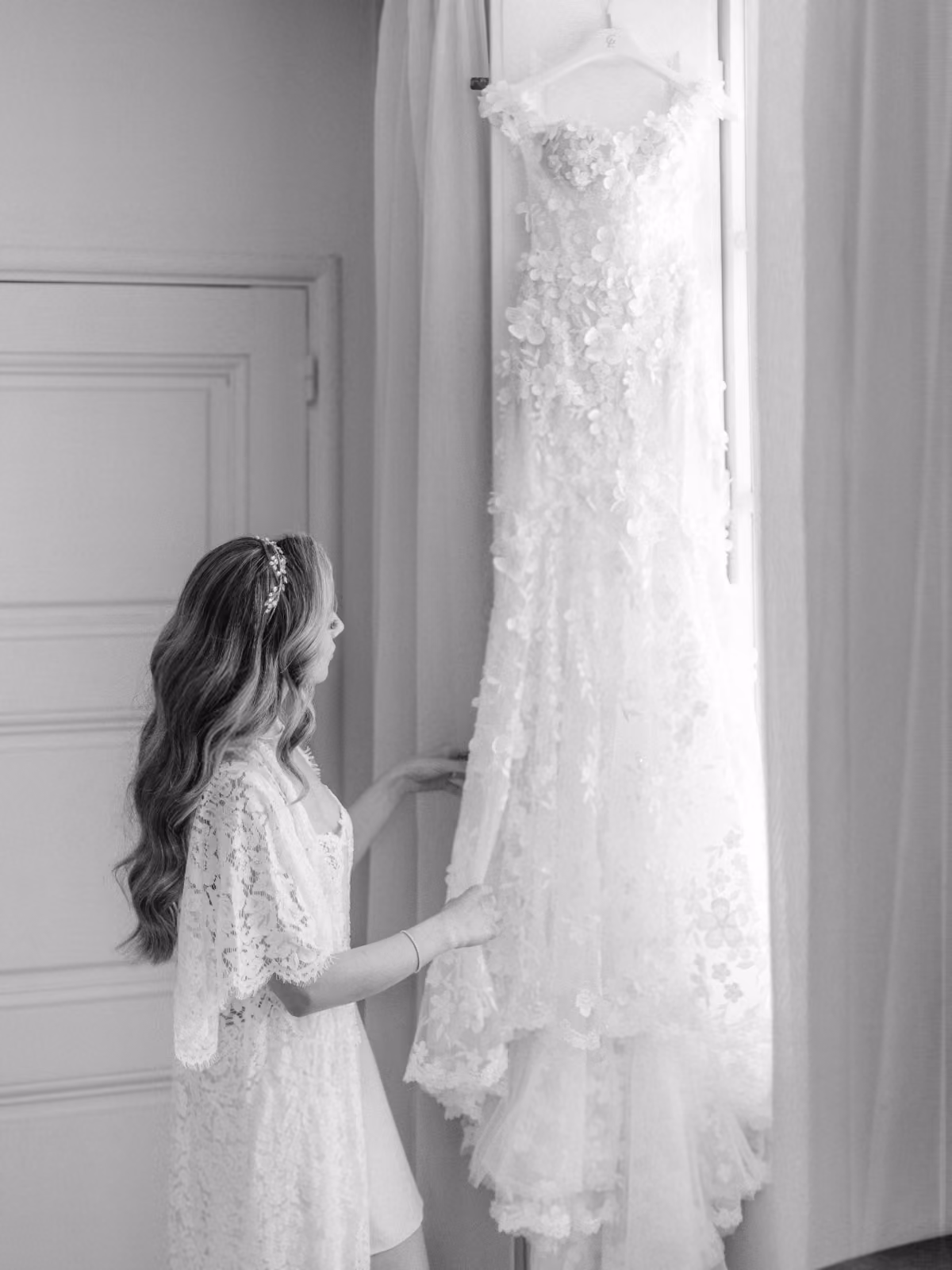 Woman in lace robe holding a floral wedding dress hanging by a window in a softly lit room.