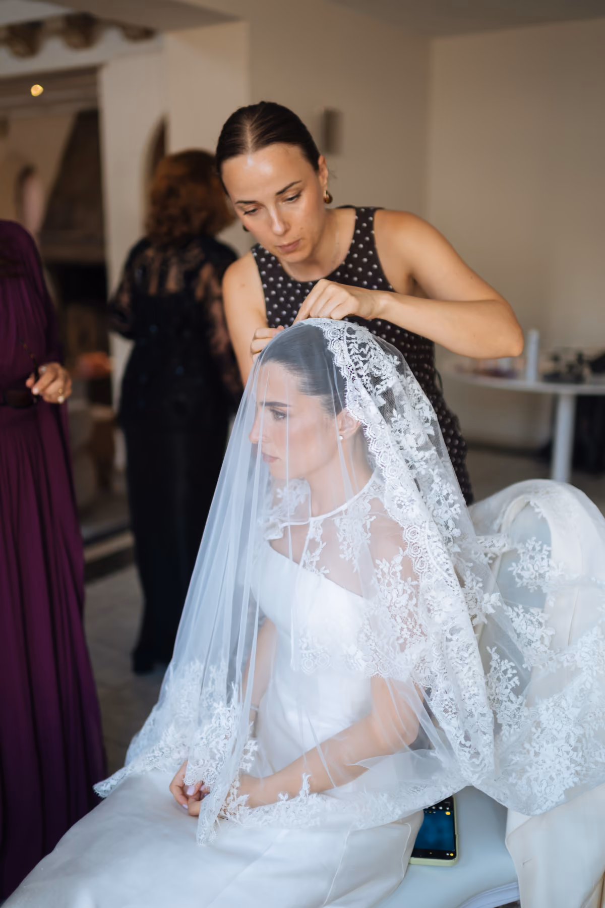 A woman adjusts the veil on a bride wearing a white wedding dress with lace details.