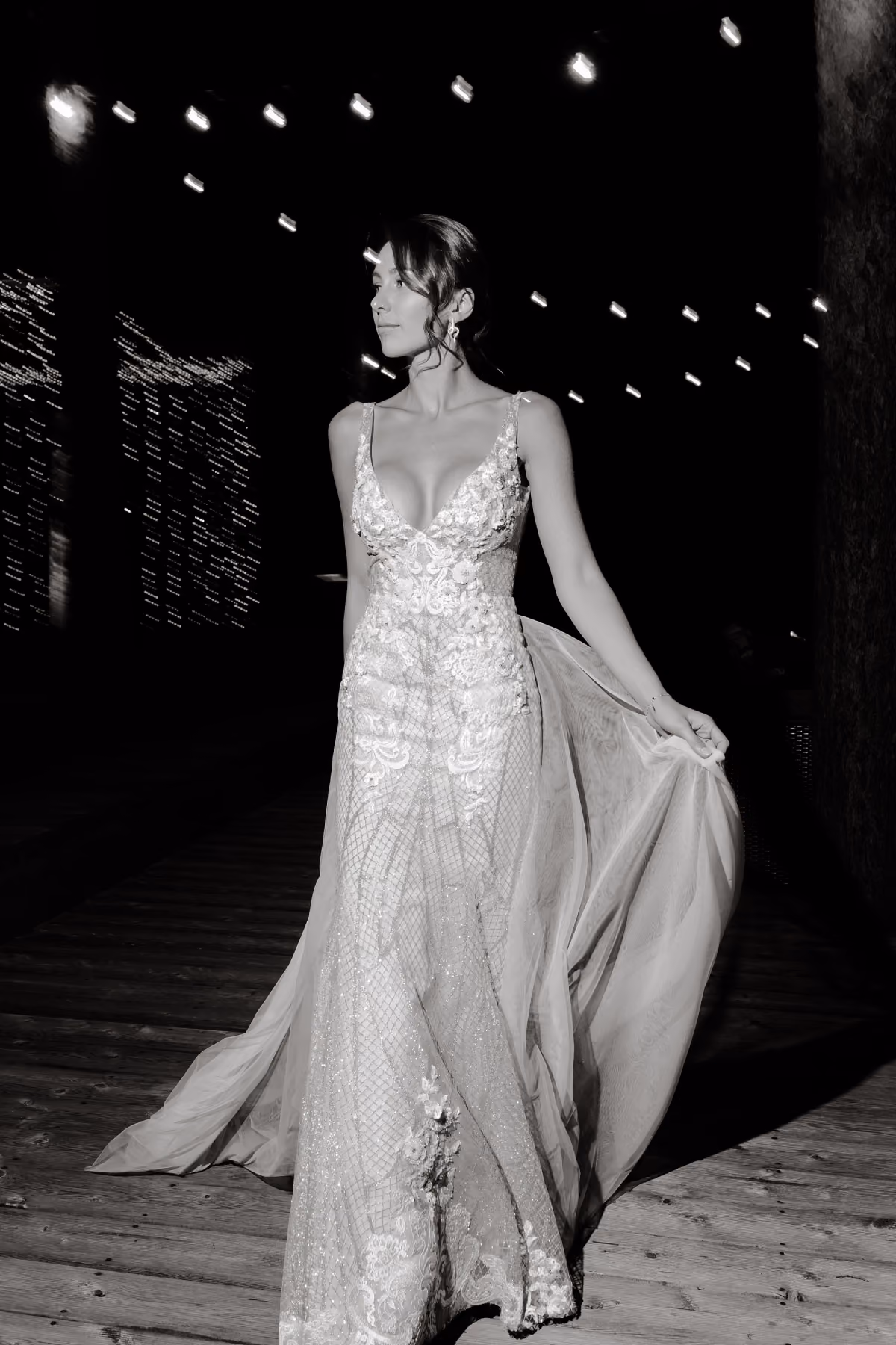 Woman in a detailed, embellished bridal gown walking on wooden floor with lights overhead at night.