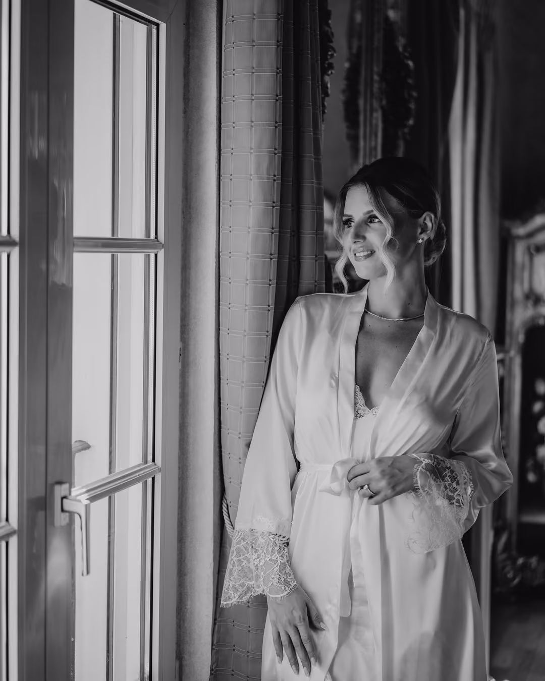 Smiling woman in a lace-trimmed robe standing by a window with curtains, looking outside.