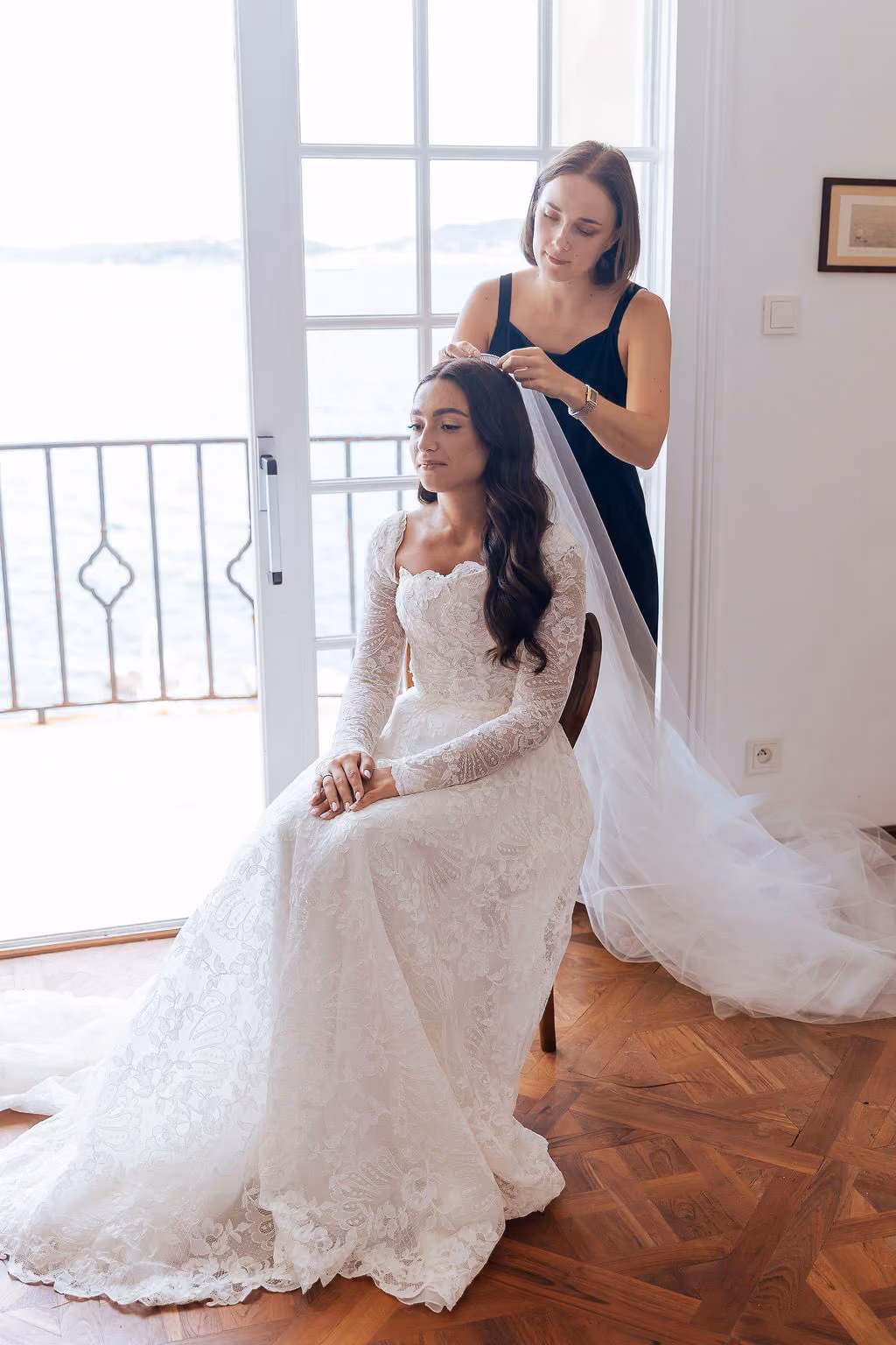 Bride in white lace gown sitting as another woman adjusts her veil near a window with a balcony view.