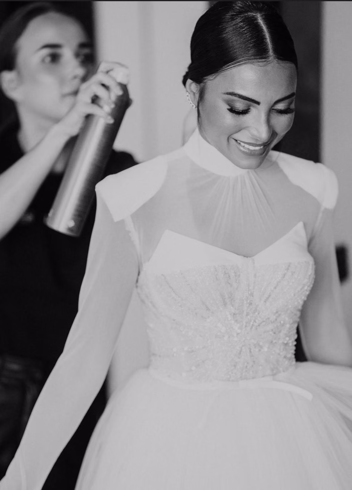 A woman in a beaded bridal gown smiles while another sprays hairspray on her hair.