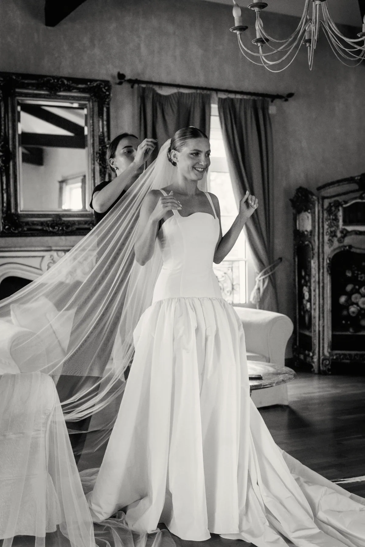 Bride smiling as another woman adjusts her long veil in an elegant room with a chandelier and curtains.