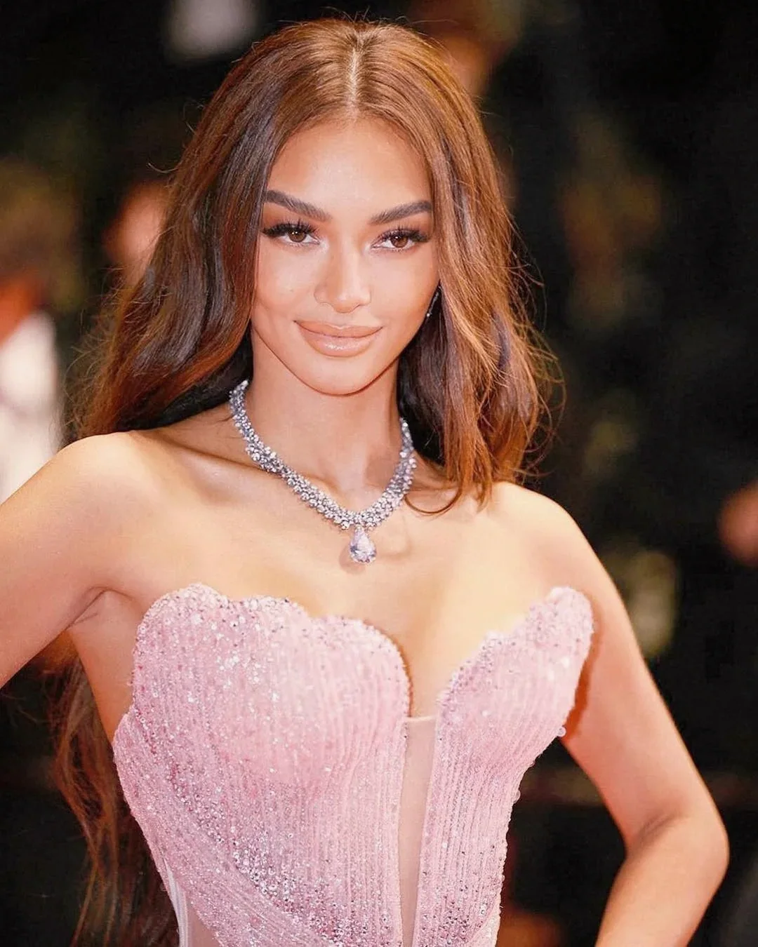 Woman with long brown hair wearing a sparkling pink strapless dress and a diamond necklace.