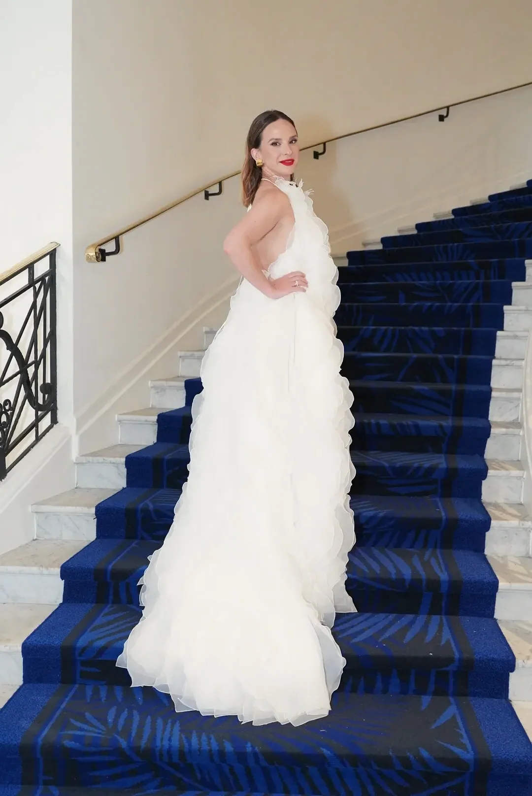 Woman in a voluminous white gown with ruffles poses on a blue-carpeted staircase.