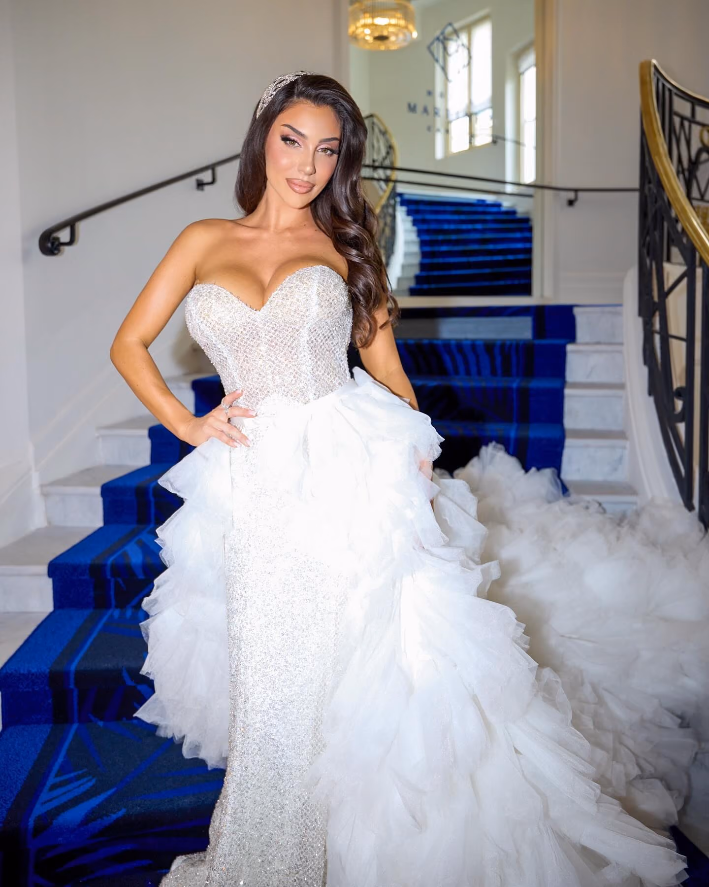 Woman in a sparkling strapless white gown with a ruffled train standing on a blue-carpeted staircase.