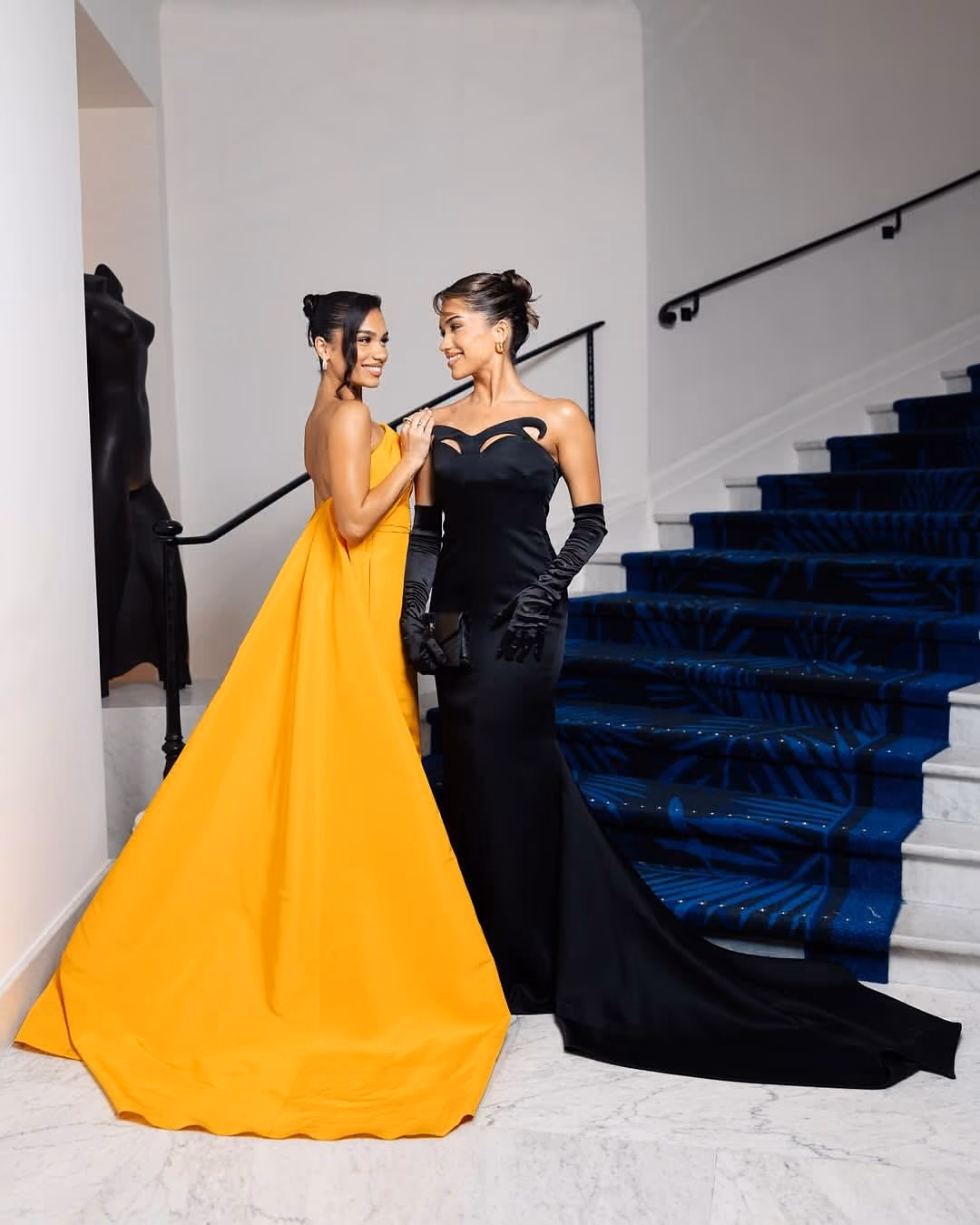 Two women in elegant gowns: one in a flowing yellow dress and the other in a fitted black dress with gloves.