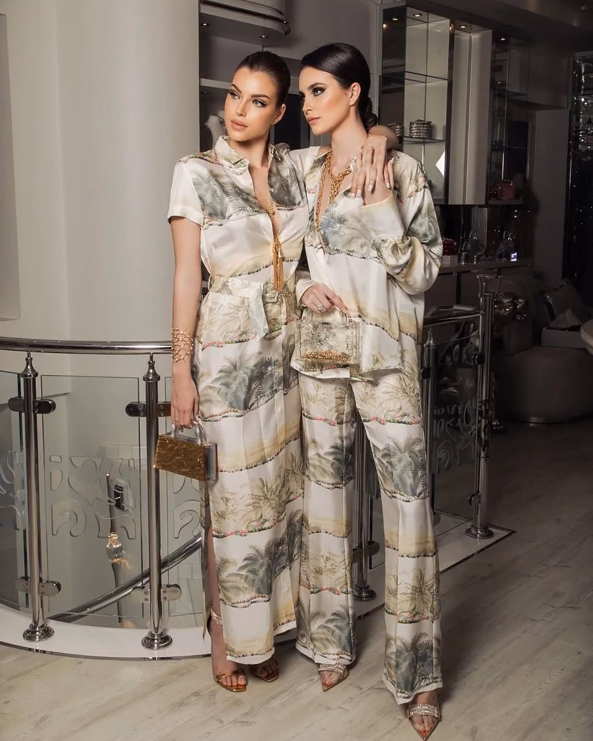 Two women in matching printed silk outfits holding small handbags inside a modern interior.