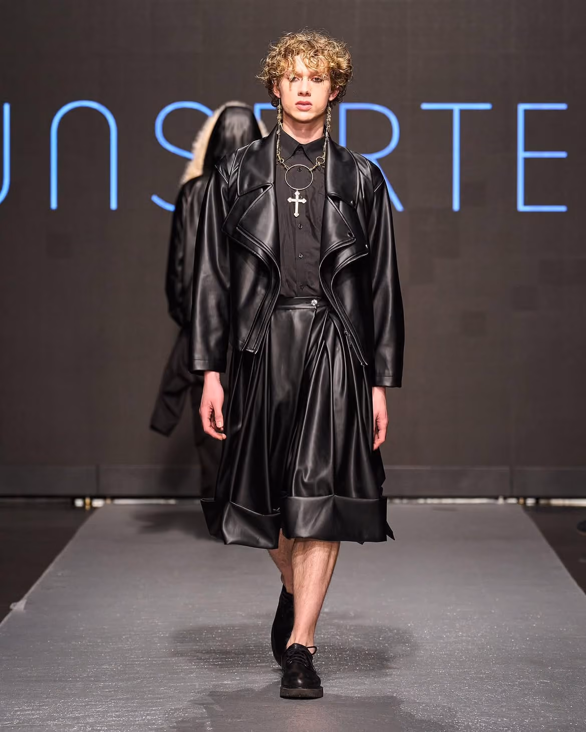 Model walking runway in black leather jacket, skirt, black shirt, and cross necklace.
