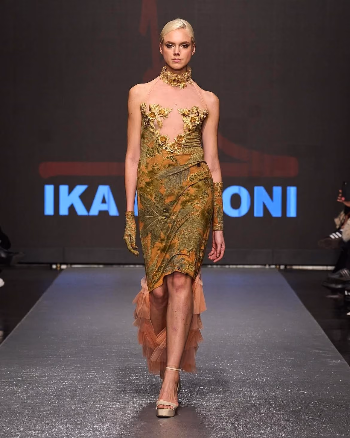 Model walking runway in green and gold floral dress with sheer neck and gloves, and ruffled back hem.