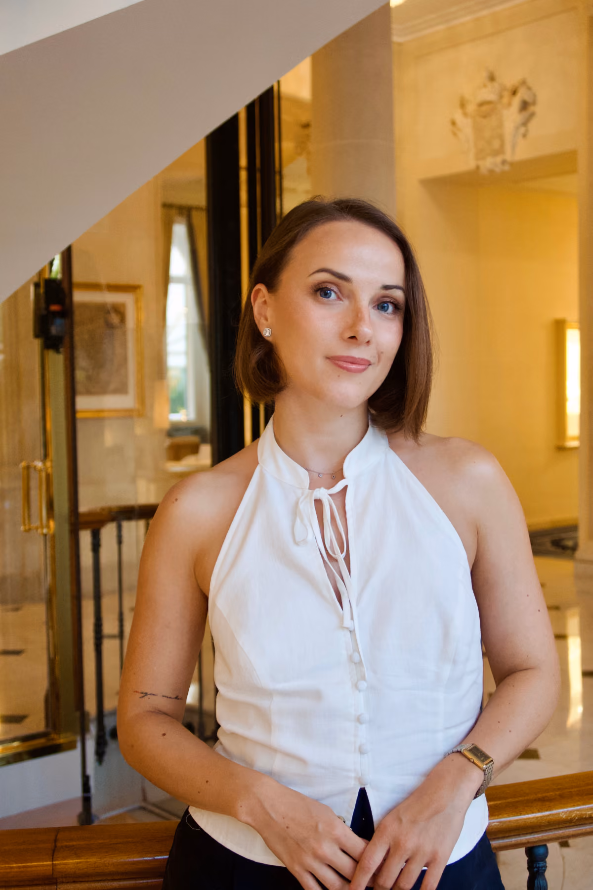 Smiling woman with short brown hair in a white sleeveless top standing indoors near a railing.