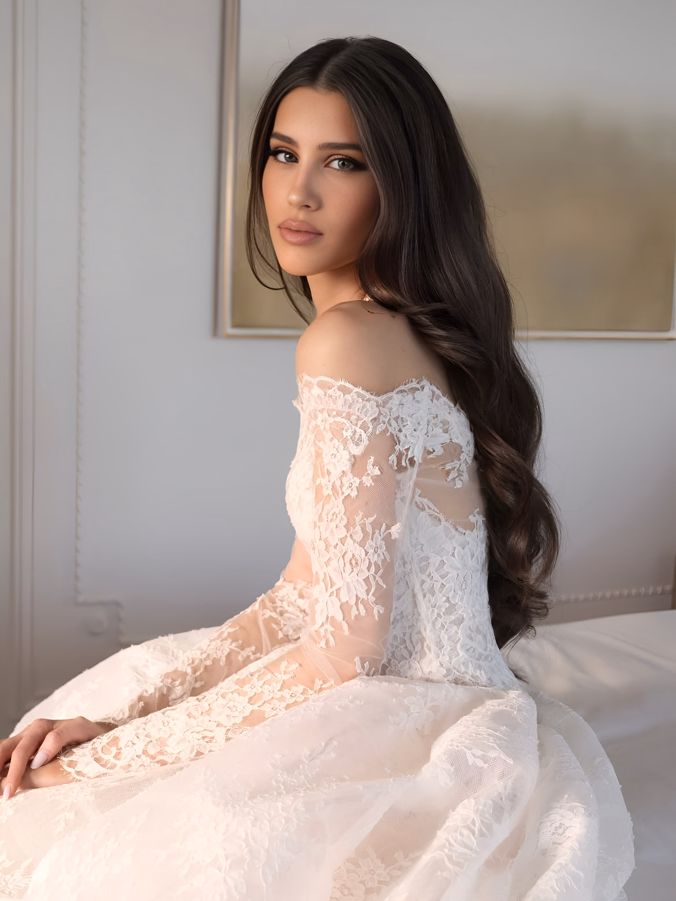 Woman with long dark hair in an off-shoulder white lace bridal gown sitting sideways indoors.