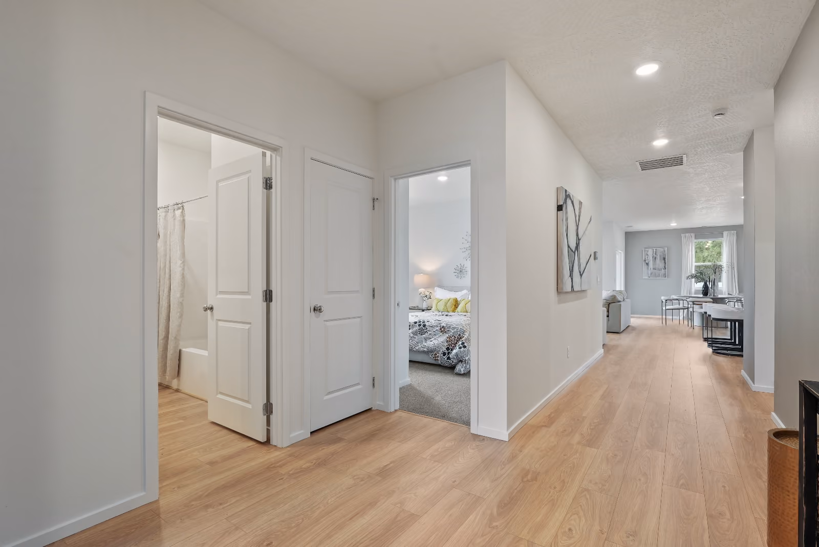The main hall in light oak flooring, looking past the second bath and a bedroom toward the living and dining room.