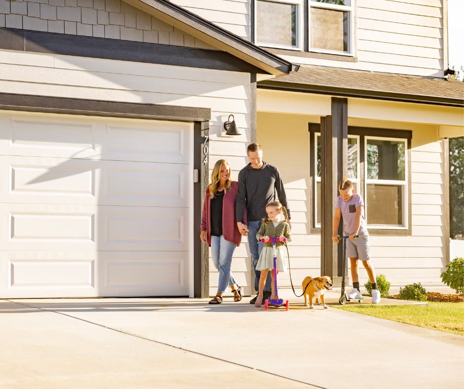 A family stepping out of their garage on a sunny afternoon, two children on scooters and a dog on a lead.