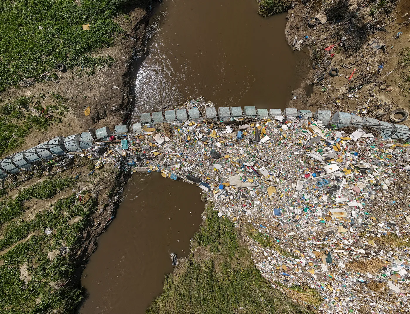 Aerial view of trash and debris collected behind a barrier in a muddy river channel.