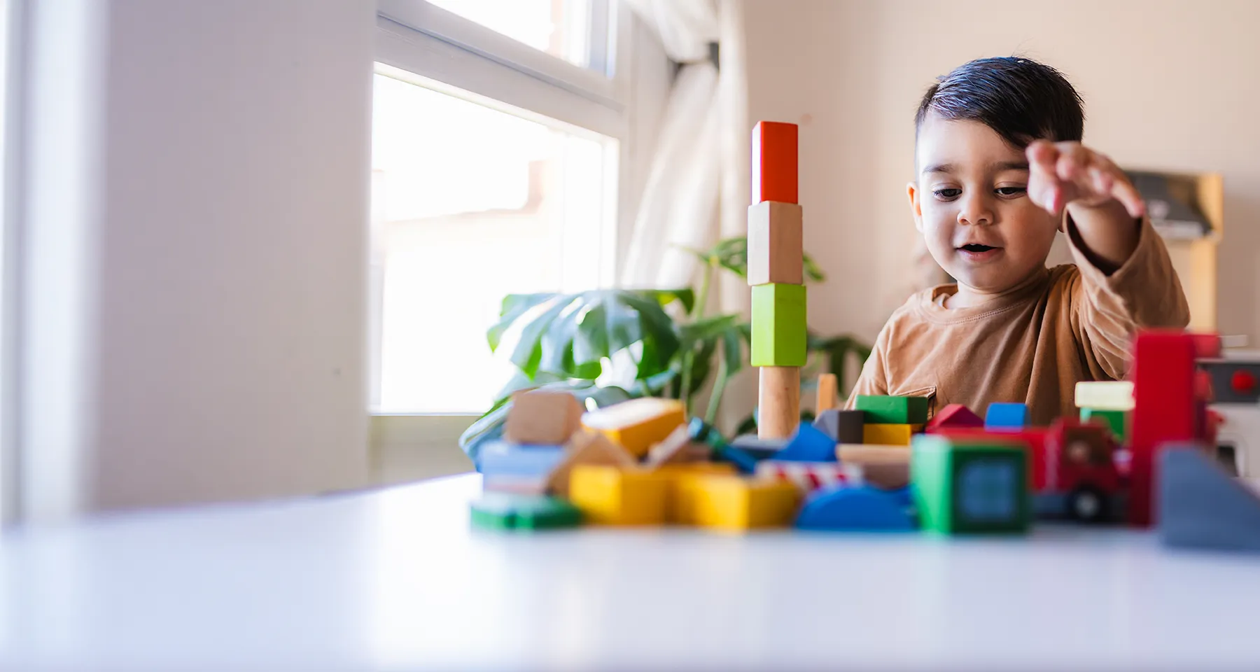 Young child builds with colorful wooden blocks at a table near a bright window.