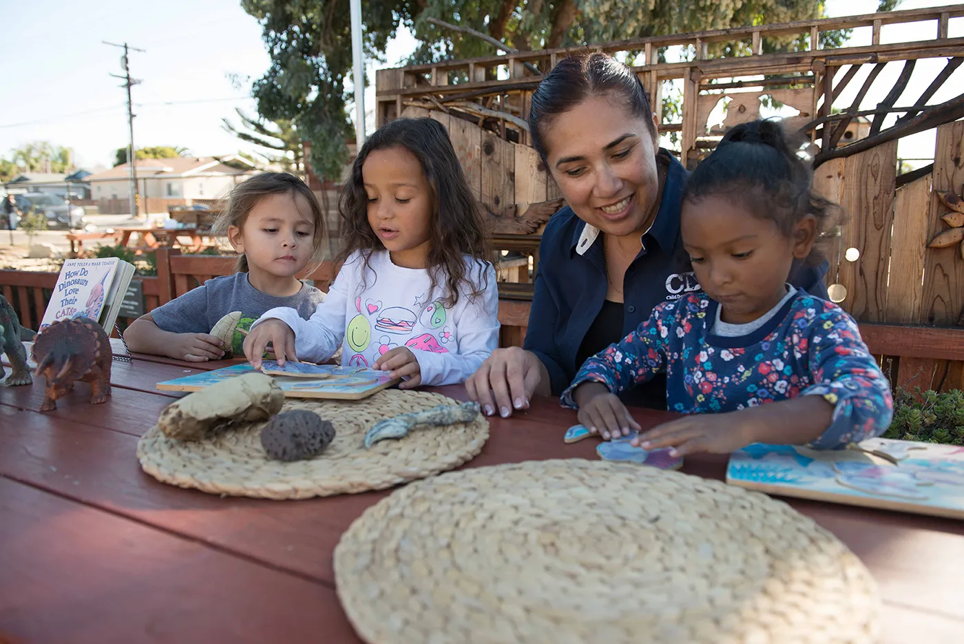 Childcare provider guides three young children through a hands-on activity at an outdoor table.
