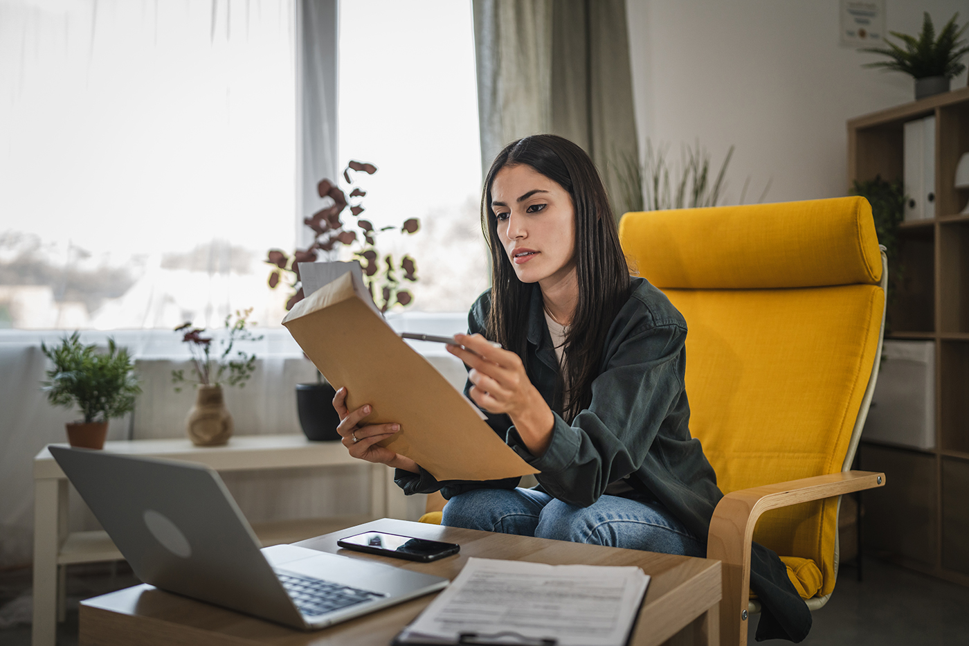 Woman reviews paperwork beside a laptop while sitting in a chair at home.