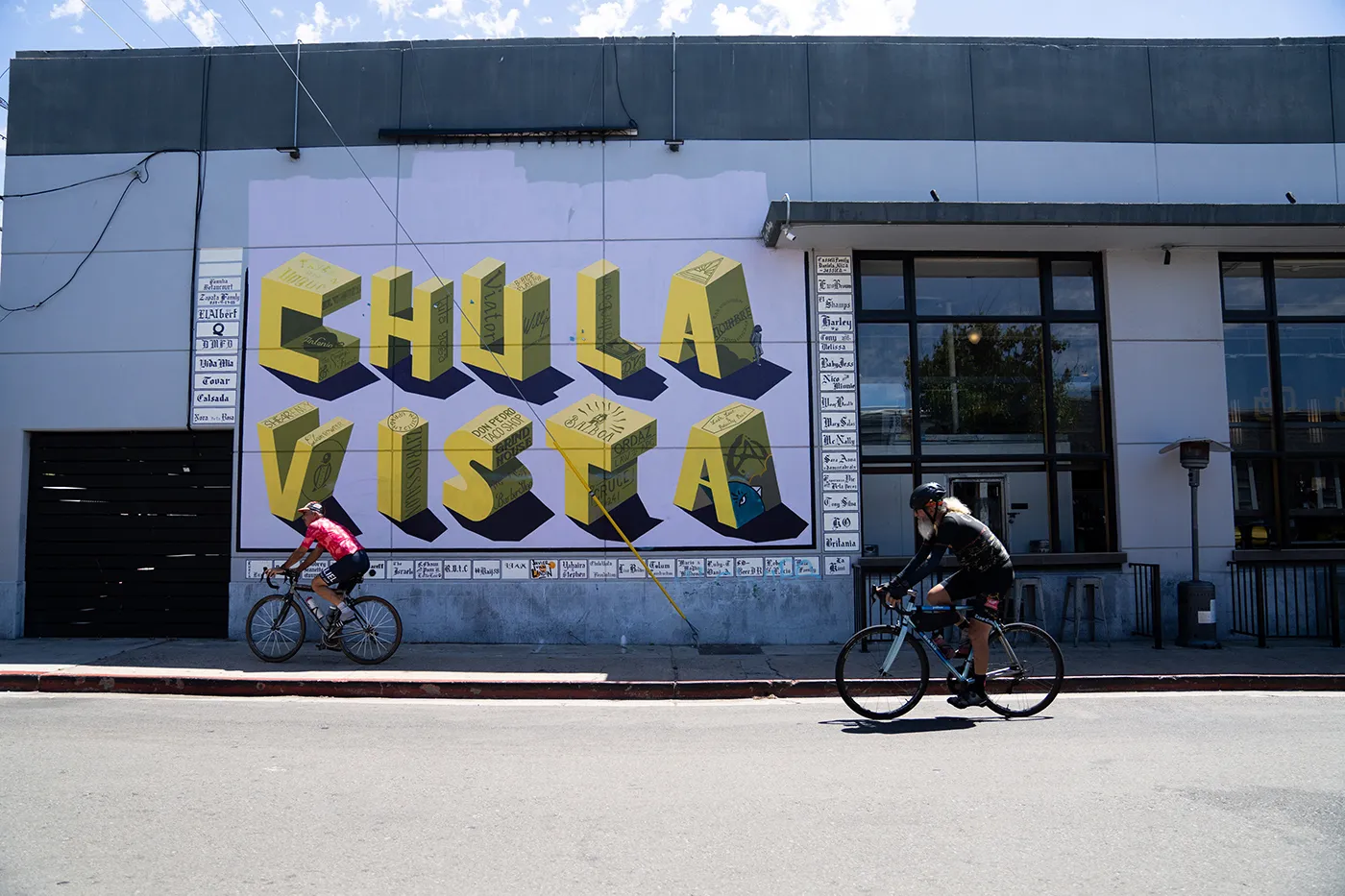 Cyclists pass a building mural spelling “Chula Vista” in large colorful letters.